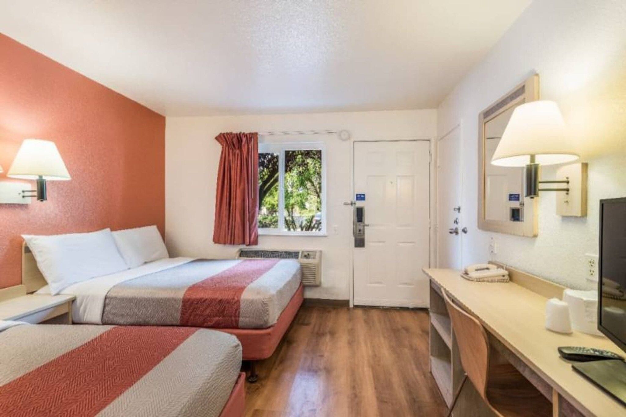 A hotel room with two beds, a desk, a mirror, a window with curtains, and a door.