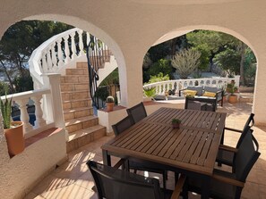 Eine überdachte Terrasse mit Holztisch und -stühlen, einer weißen Treppe und Blumentöpfen.