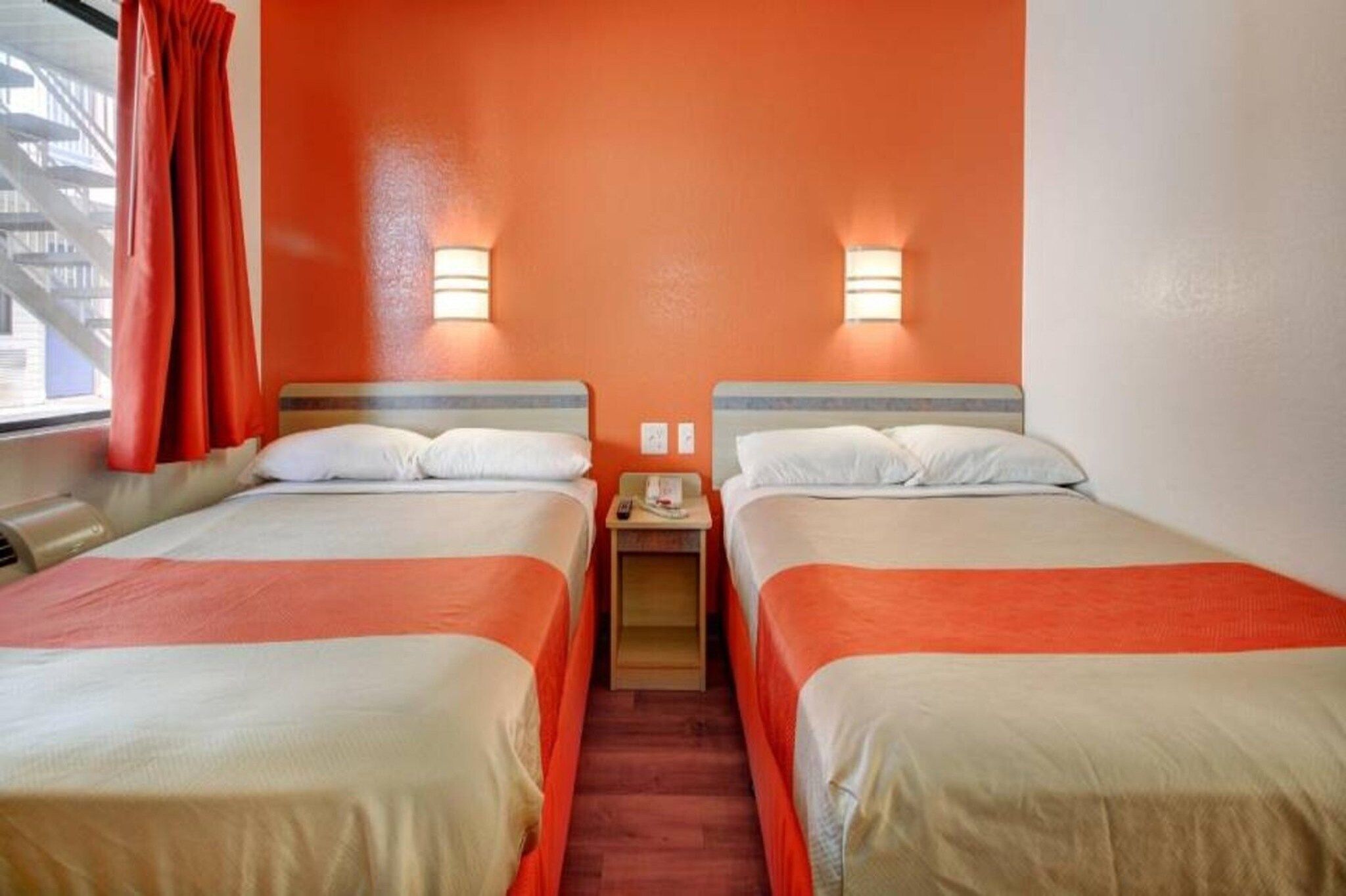 A hotel room with two beds, a window with red curtains, and a small table with a lamp.