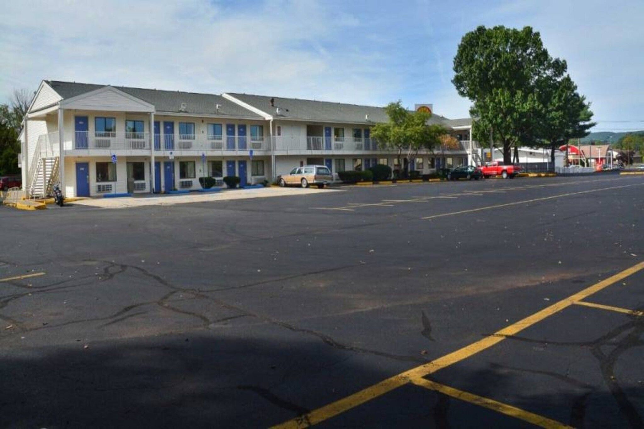 Un motel con una hilera de habitaciones, un estacionamiento y un árbol grande al fondo.