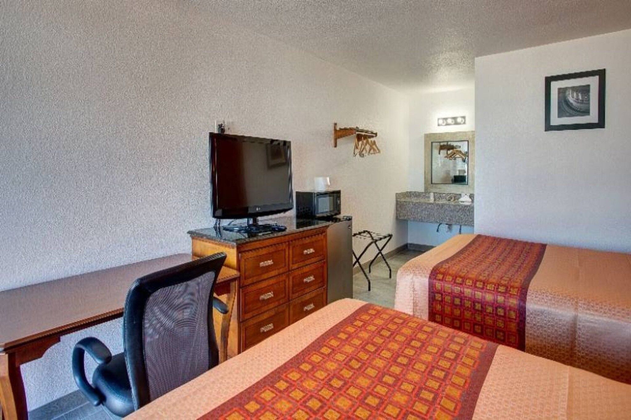 A hotel room with a desk, a television, a microwave, a small bathroom with a mirror, and two beds with patterned bedspreads.