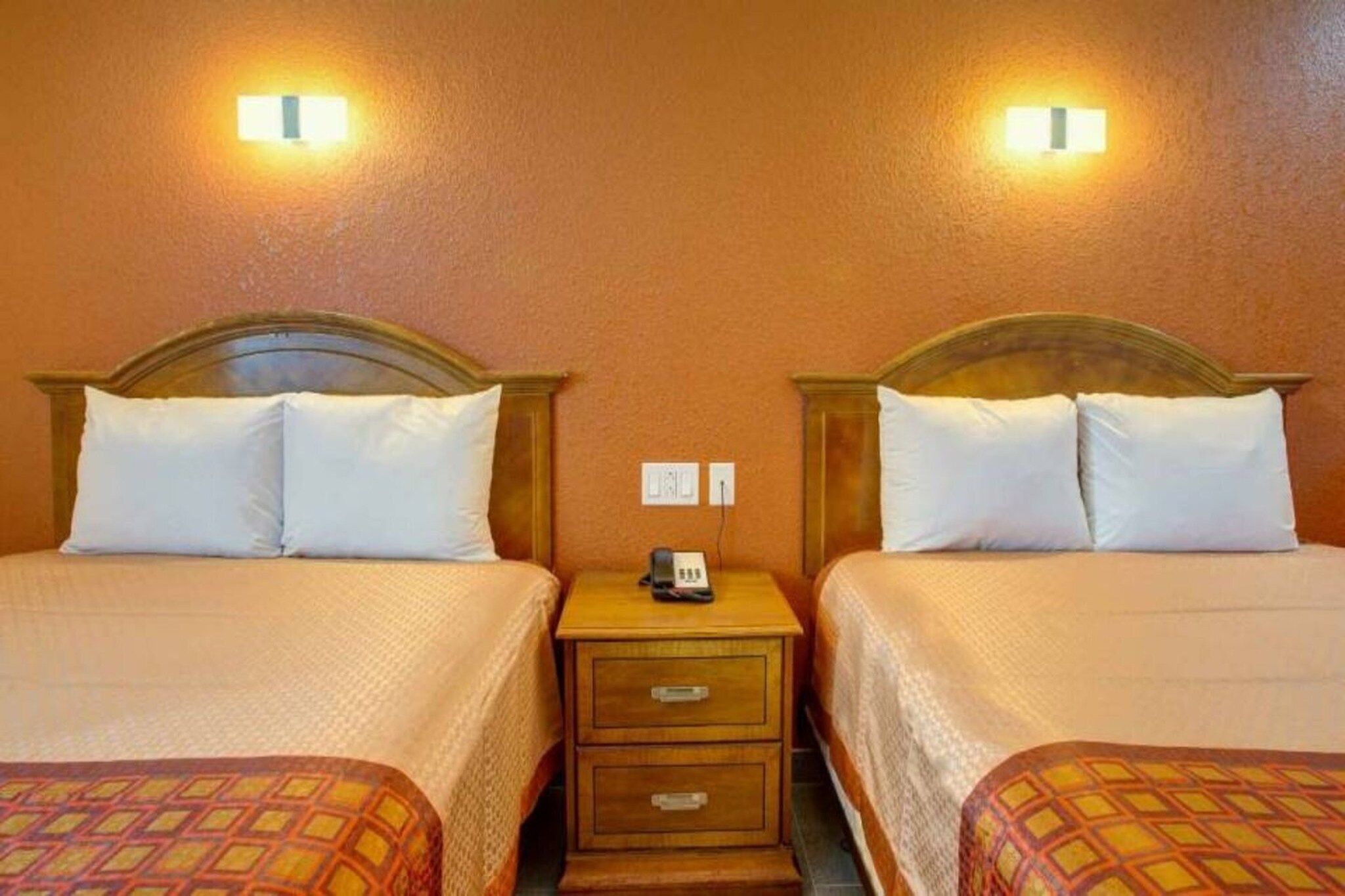 A hotel room with two beds, each with a headboard, a bedside table with a phone, and a wall socket.