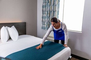 A hotel room with a bed, pillows, a blue bedspread, and a person adjusting the bedspread. - 1 King Bed | Non-Smoking, Micfridge (Branson MO)