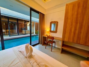 A hotel room with a bed, a desk, a chair, a wooden wardrobe, a large window with a view of a swimming pool, and a wall-mounted mirror. - Villa Sora by Kelle, Ubud (Gianyar)