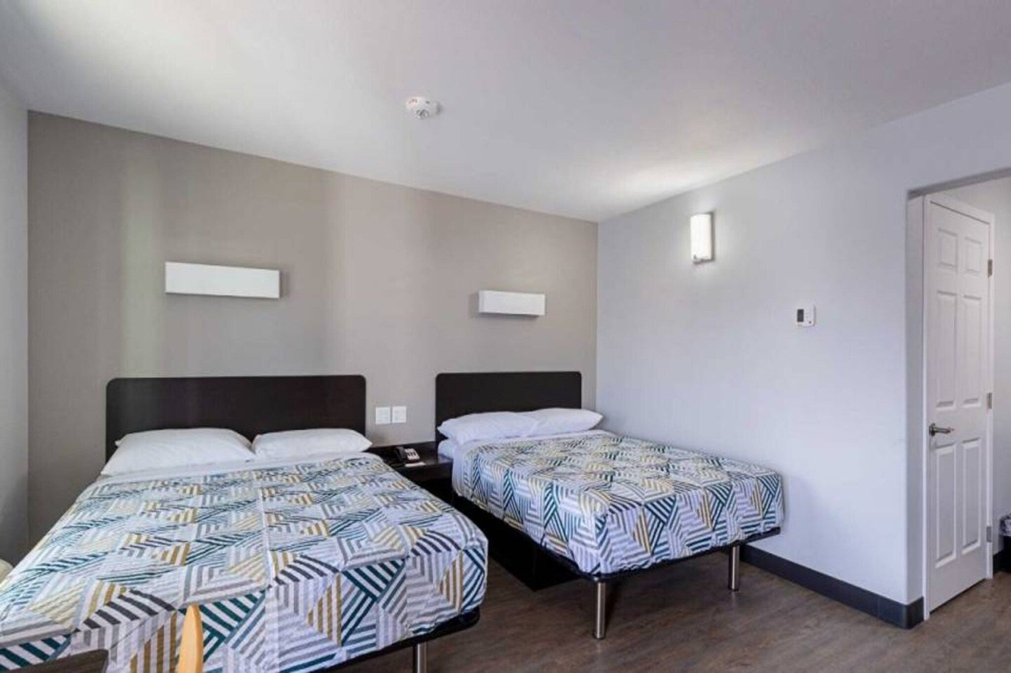 Una habitación de hotel con dos camas individuales, una mesita de noche y una lámpara de pared, además de una puerta.