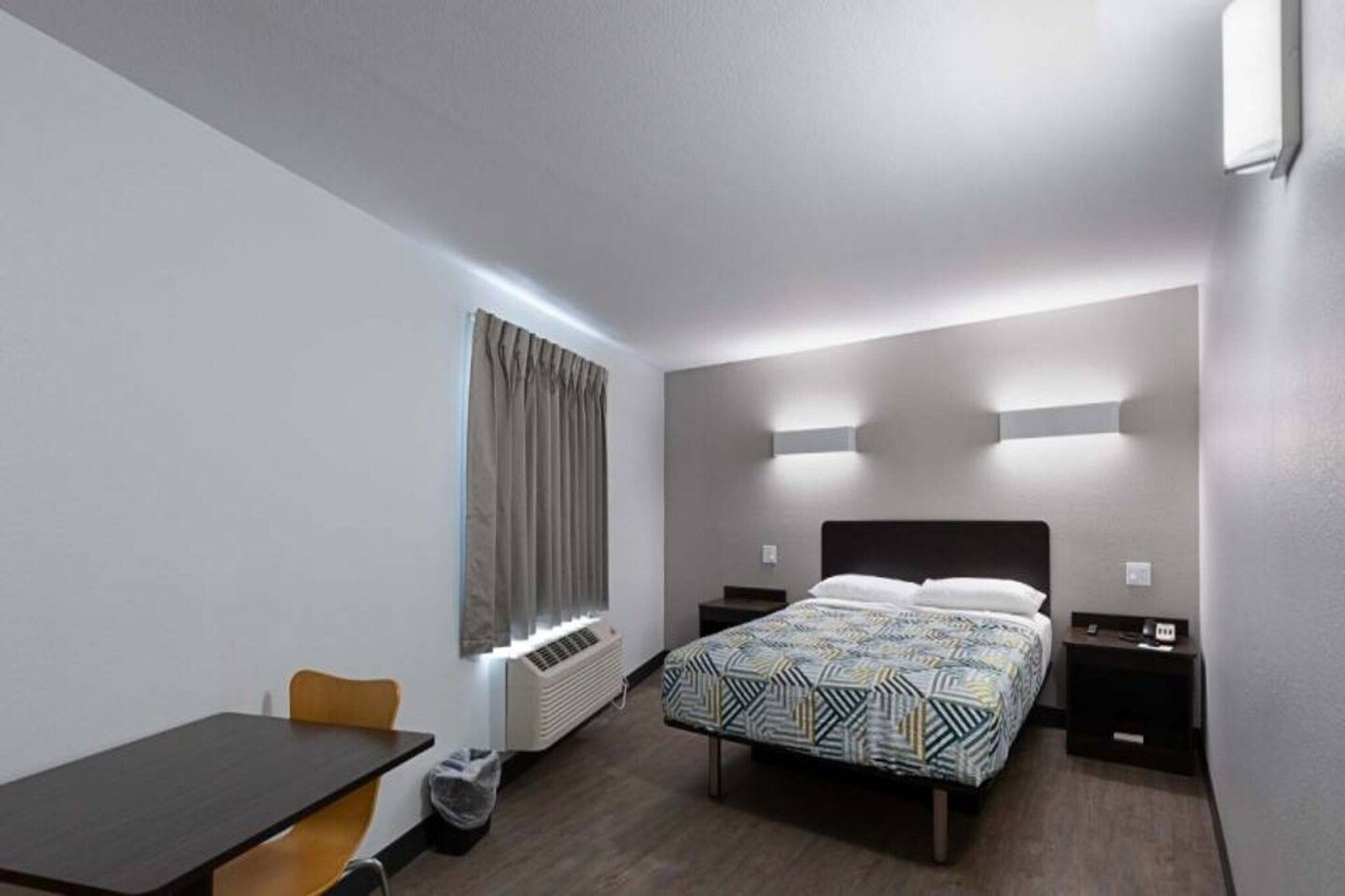 A hotel room with a bed, bedside tables, a desk, a chair, a window with curtains, and a wall-mounted light fixture.