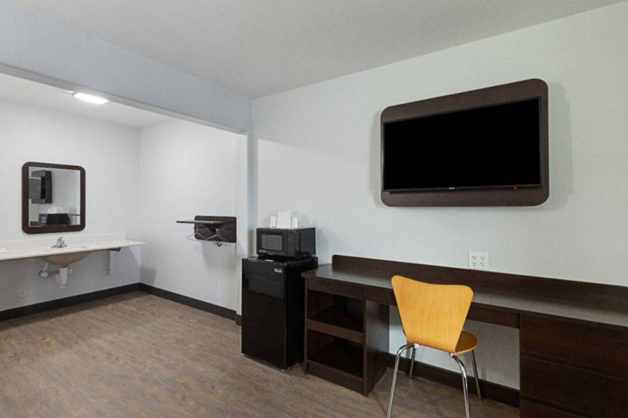 A hotel room with a flat-screen TV mounted on the wall, a small desk with a chair, a sink, and a mirror.