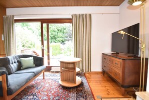 Ein Wohnzimmer mit einer Couch, einem runden Holztisch, einem Fernseher auf einer Holzkommode und einem großen Fenster mit Vorhängen.