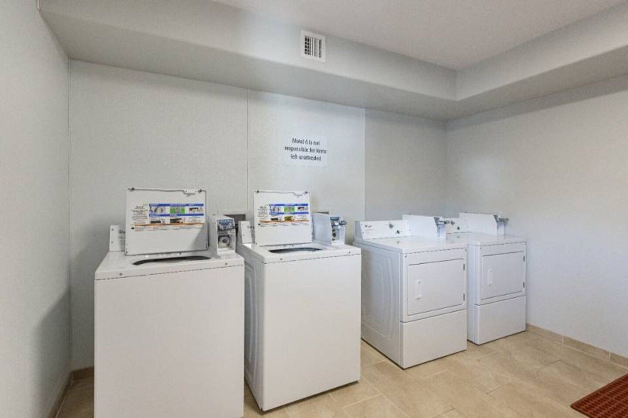 A laundry room with four white washing machines and dryers, a vent on the ceiling, and a sign on the wall.