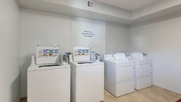 A laundry room with four white washing machines and dryers, a vent on the ceiling, and a sign on the wall.