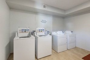 Une buanderie équipée de quatre machines à laver et de quatre sèche-linge blancs, avec une bouche d’aération au plafond et un panneau affiché sur le mur.