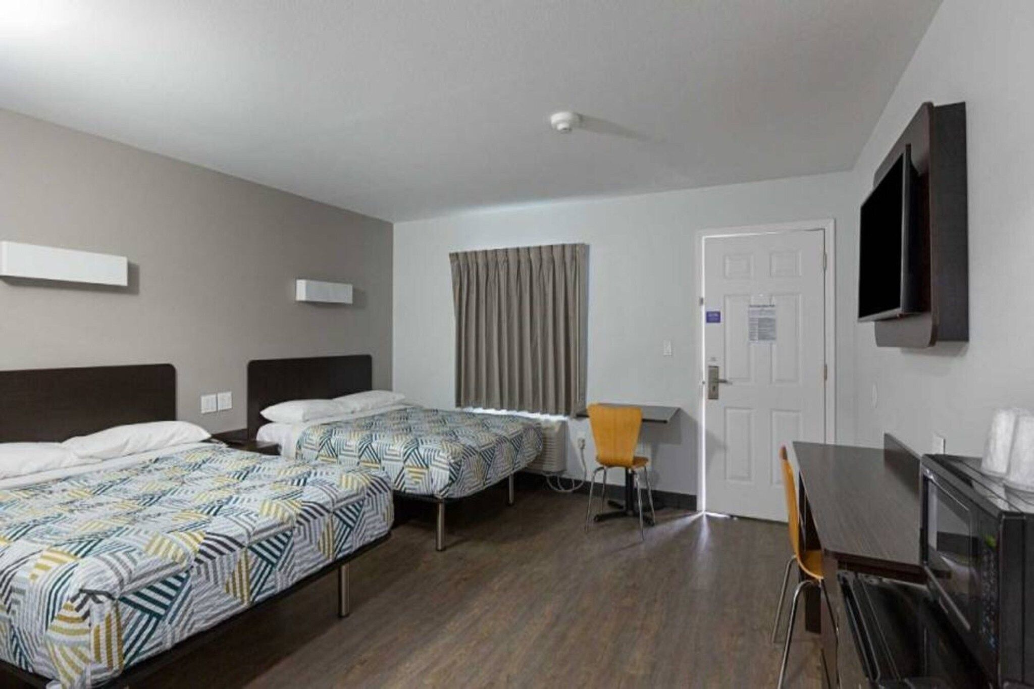 A hotel room with two single beds, a desk, a chair, a TV mounted on the wall, and a door.