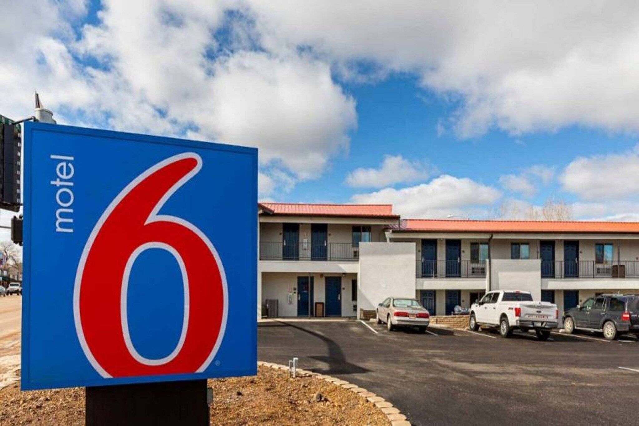 A motel sign with the number 6, a parking lot, and a building with multiple windows and a red roof.