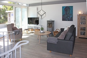 A modern living room with a grey sofa, a round coffee table, and a TV. There is a painting on the wall and a cabinet with glass doors. - fewo1846 - StrandNest - House with a view of the fjord, a fireplace, a sauna, and 3 bedrooms - (Glücksburg (Ostsee))