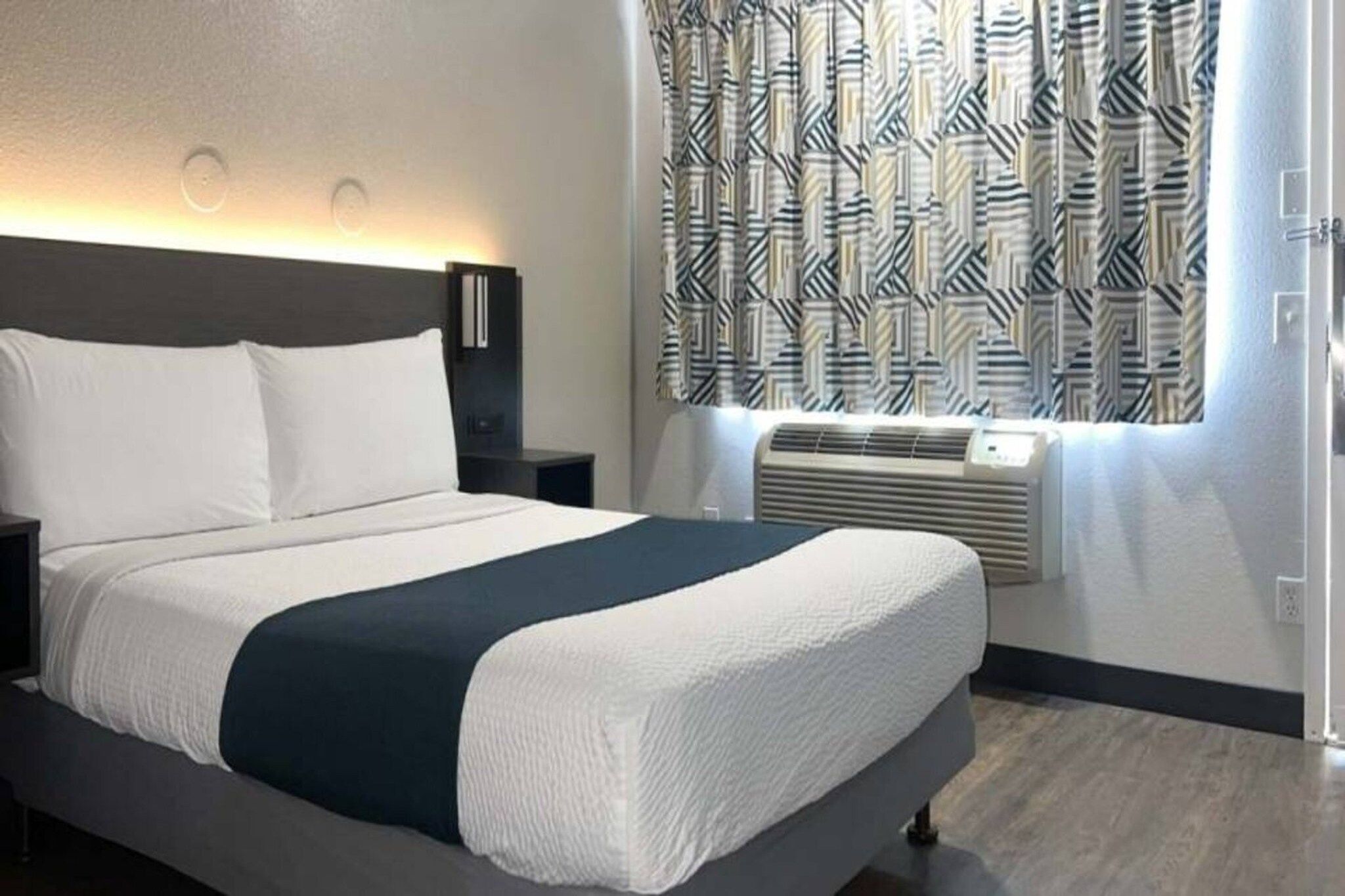 A hotel room with a bed, bedside tables, a wall-mounted heater, and patterned curtains.