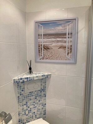 A bathroom with a tiled wall, a shower area, and a framed picture of a beach scene. - Ankerplatz - Your cozy anchorage in Westerland on Sylt for a (Sylt)