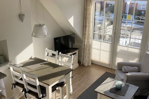 A room with a dining table, chairs, a television, a sofa, and a view of the outside. - Ankerplatz - Your cozy anchorage in Westerland on Sylt for a (Sylt)