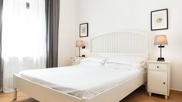 Een wit bed met hoofdeinde, twee nachtkastjes met lampen, een raam met gordijnen en twee ingelijste foto's aan de muur.