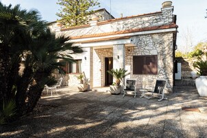 Un edificio in pietra con tetto a tegole, una porta e una finestra con persiane. Sono presenti piante in vaso e mobili da esterno.