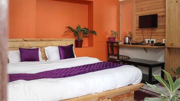 Un dormitorio con cabecera de madera, cama con ropa de cama blanca y morada, una mesita de madera, una silla, un televisor montado en la pared y una planta en maceta.