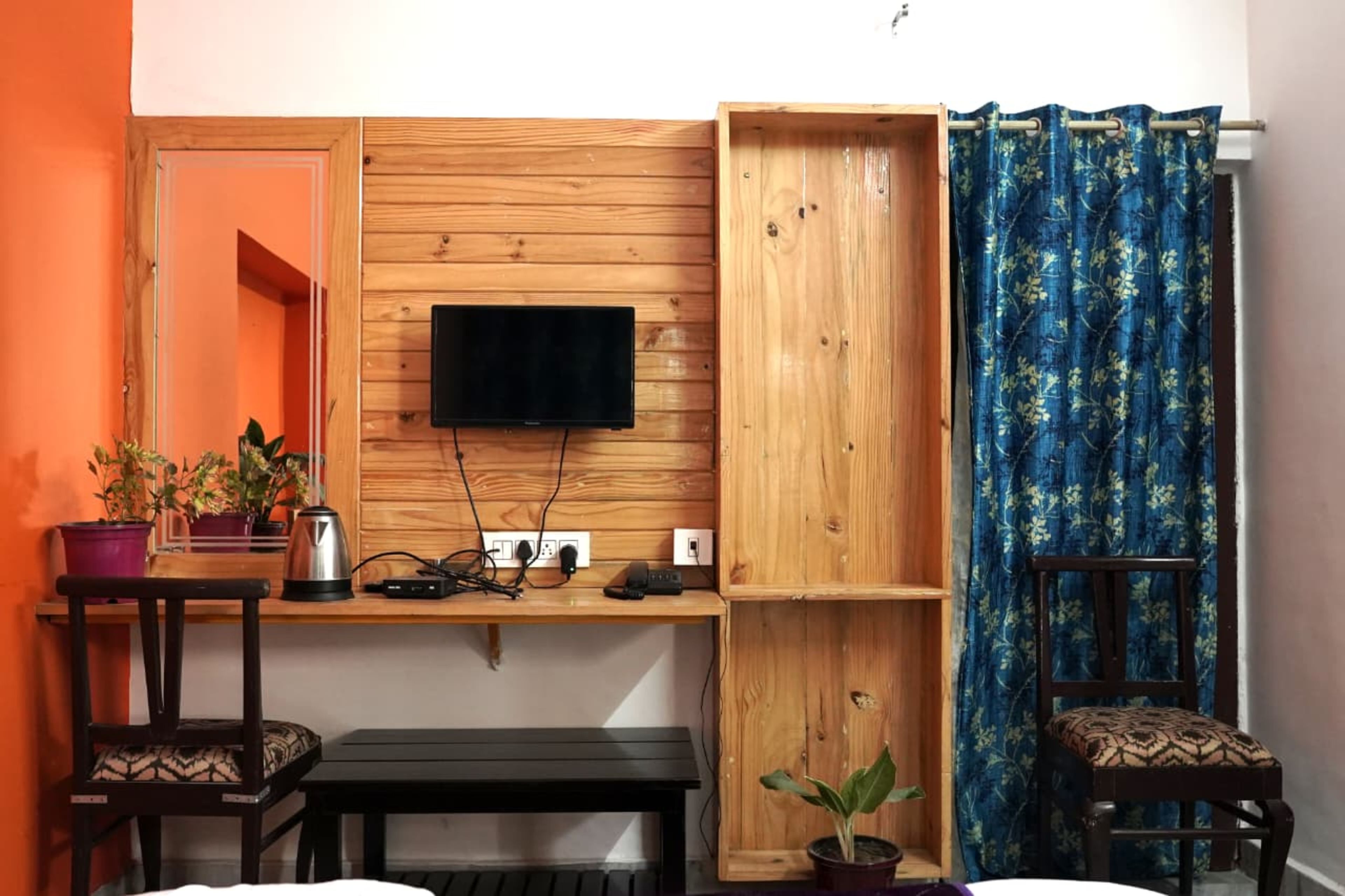 Une pièce de taille modeste, avec un téléviseur fixé au mur, un petit meuble, une bouilloire, un banc et une chaise.