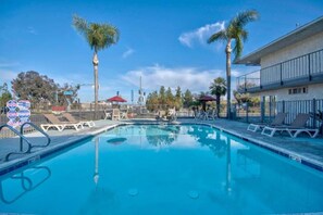 A clear blue swimming pool with lounge chairs and palm trees, surrounded by a fenced area and a building with a balcony. - Cozy Queen Bedroom|Pool|Free Wifi (Tulare)