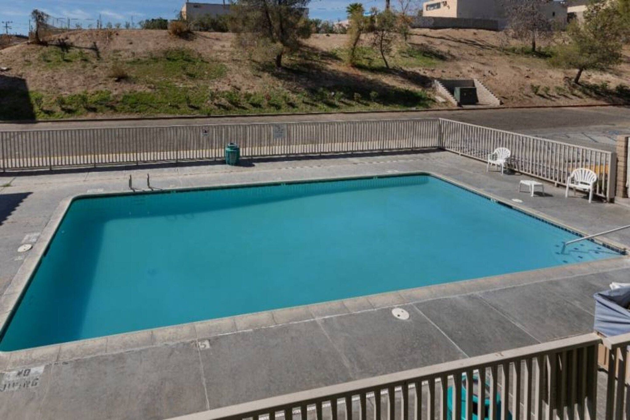 A rectangular swimming pool with a tiled deck surrounding it, a metal fence, and a few chairs and a trash can.