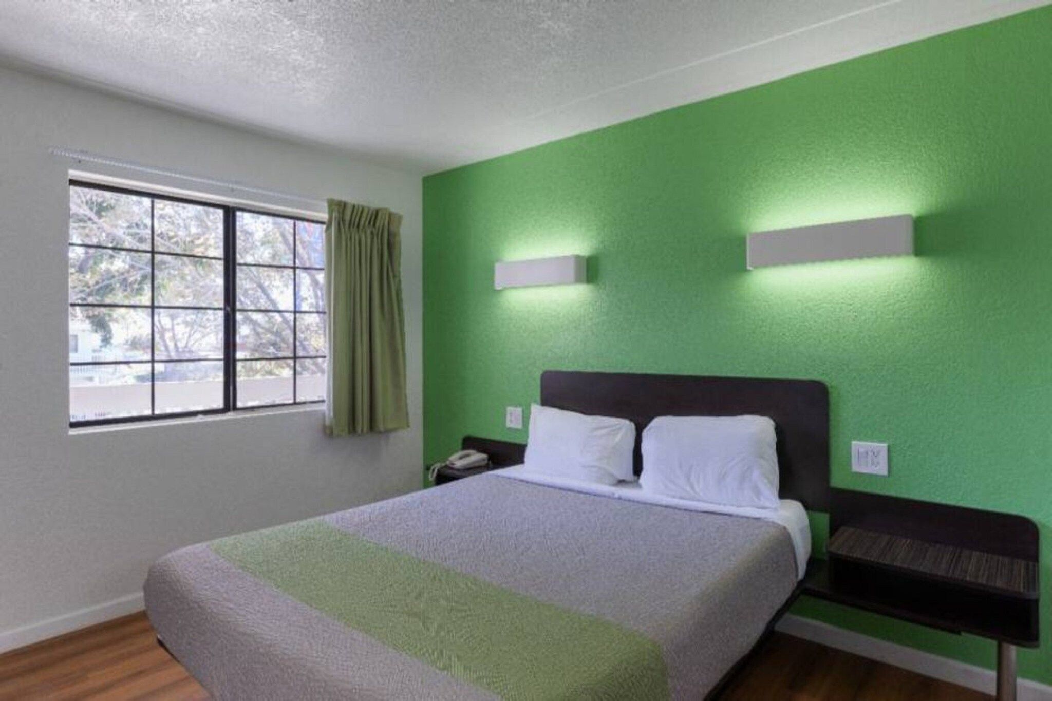 Un dormitorio con cama, mesita de noche, lámpara de pared y ventana con cortinas.