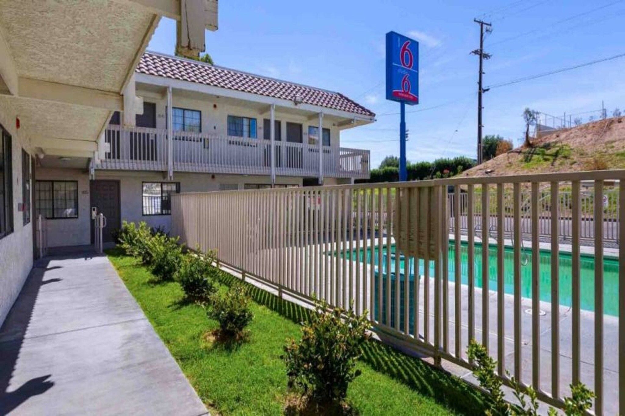 A motel with a fenced pool area, a covered parking lot, and a clear sky.