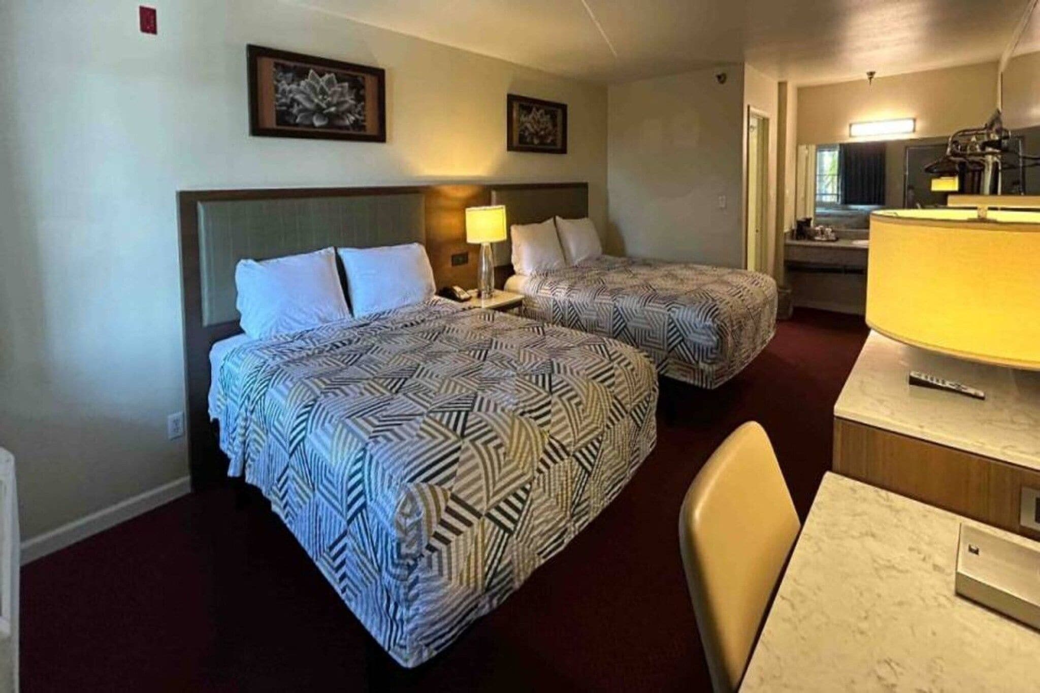 A hotel room with two beds, a desk, a chair, a lamp, and artwork on the walls.