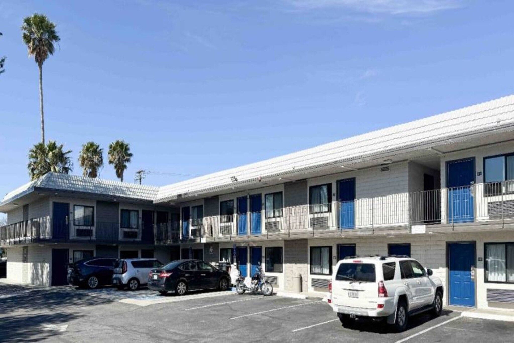 A multi-story motel with a parking lot, several cars parked, and palm trees in the background.