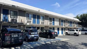 Un motel à étages, avec une rangée de chambres, chacune dotée d’un balcon. Plusieurs voitures sont garées devant, et une place de stationnement réservée aux personnes handicapées est visible.