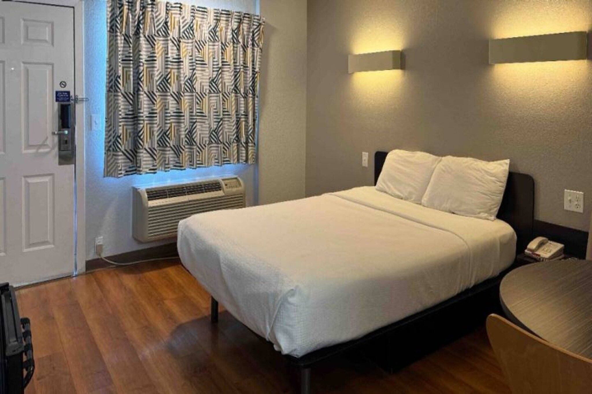 Een hotelkamer met een bed, een raam met gordijnen met een patroon, een airconditioningsunit en een nachtkastje met een telefoon.