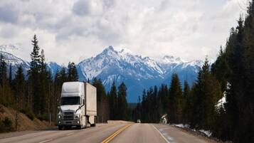 A road with a large truck and a smaller vehicle, surrounded by snow-capped mountains and dense forests.