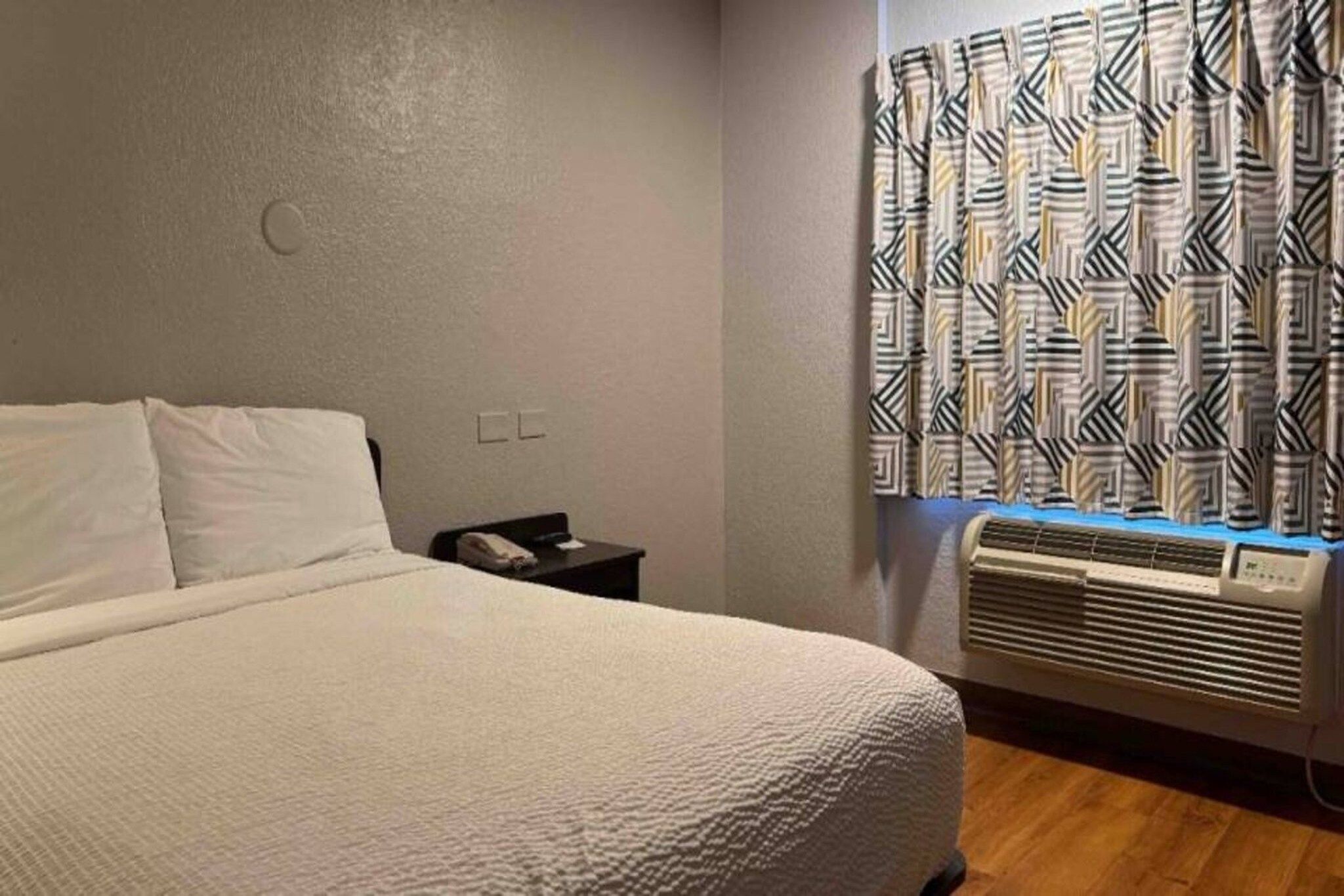 Een slaapkamer met een bed, een nachtkastje, een wandklok, een raam met gordijnen met een patroon en een luchtverfrisser aan de muur.