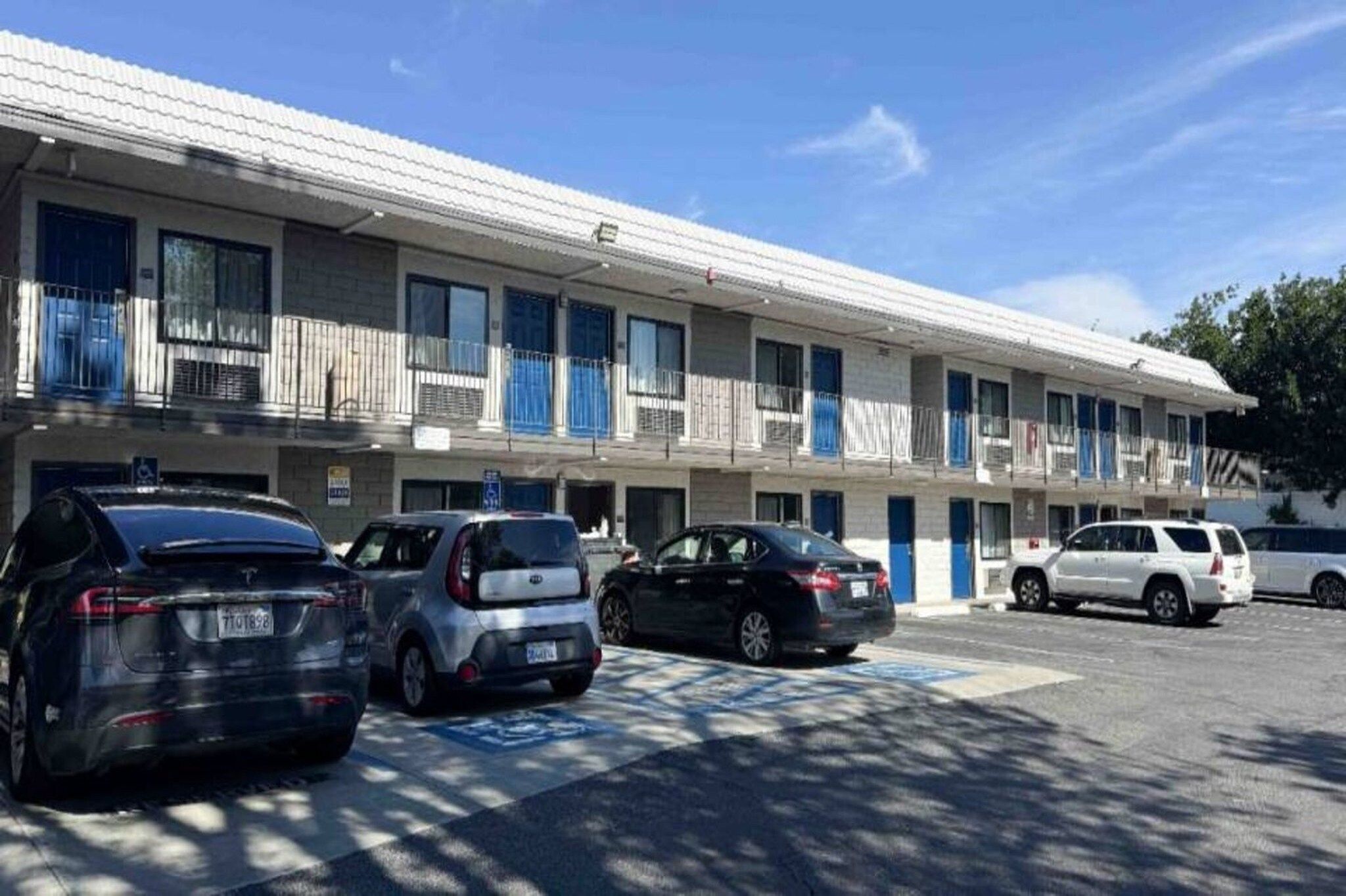 A multi-story motel with a row of rooms, each with a balcony. There are several cars parked in front, and a handicapped parking space is visible.