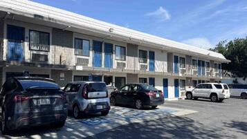 A multi-story motel with a row of rooms, each with a balcony. There are several cars parked in front, and a handicapped parking space is visible.