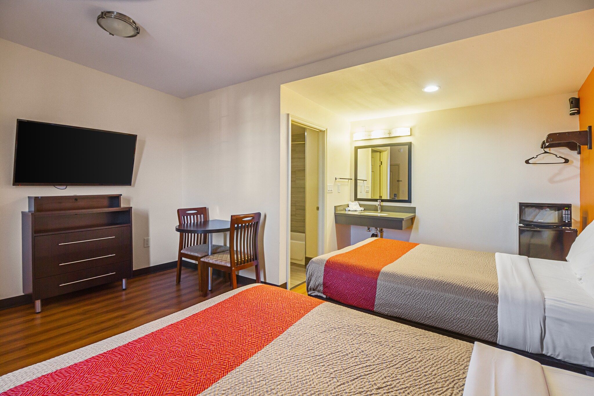 A hotel room with a TV, a desk with drawers, two chairs, a bed with a patterned blanket, a bathroom with a mirror and sink, and a microwave.