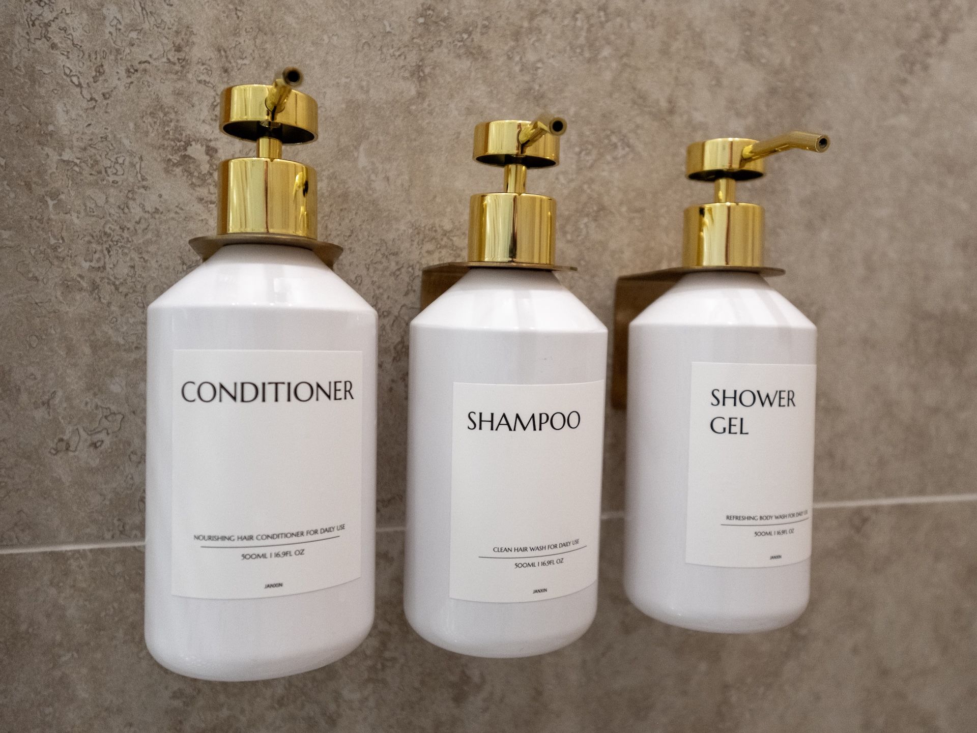 Three white bottles with gold pumps against a textured wall. Each bottle is labeled with a different product: conditioner, shampoo, and shower gel.