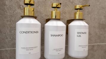 Three white bottles with gold pumps against a textured wall. Each bottle is labeled with a different product: conditioner, shampoo, and shower gel.