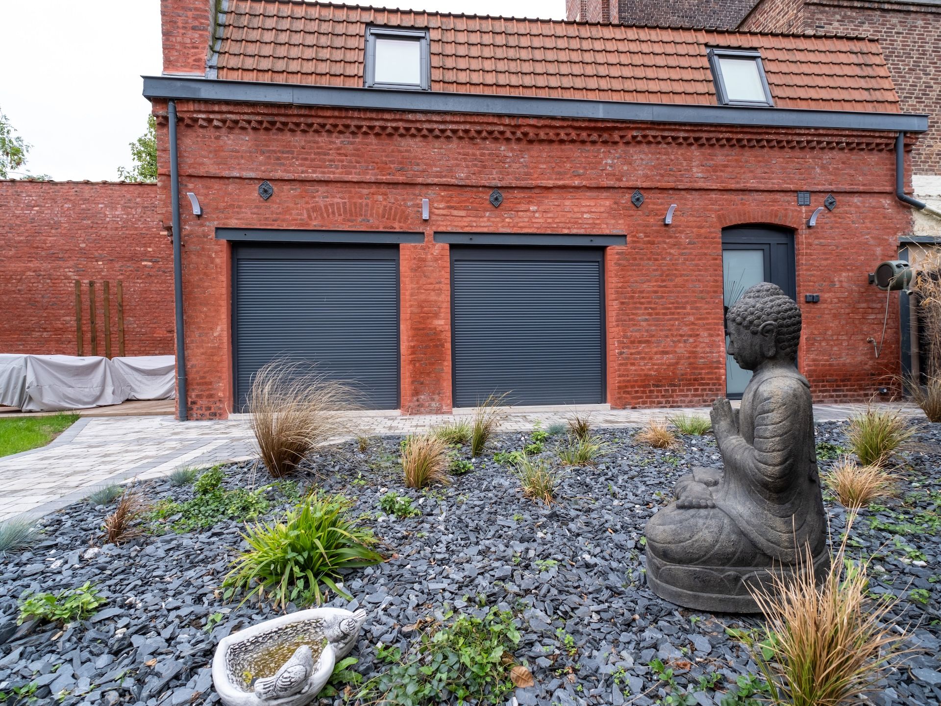 Un bâtiment à la façade en briques, avec deux portes de garage fermées et une statue de Bouddha en premier plan.