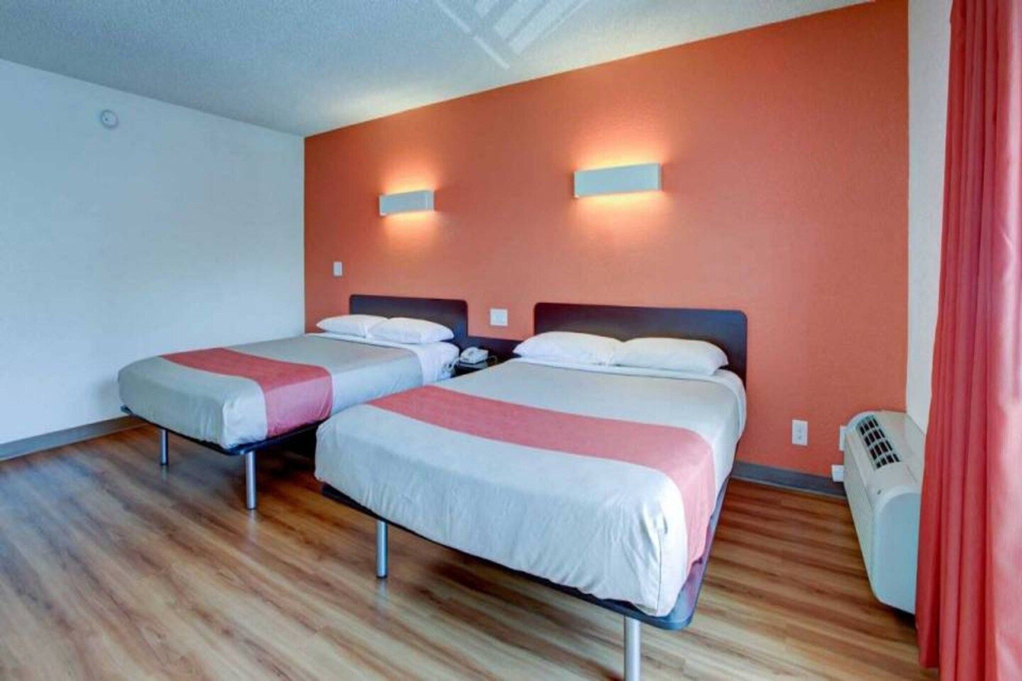 A hotel room with two beds, wooden flooring, a red curtain, and a wall-mounted heater.