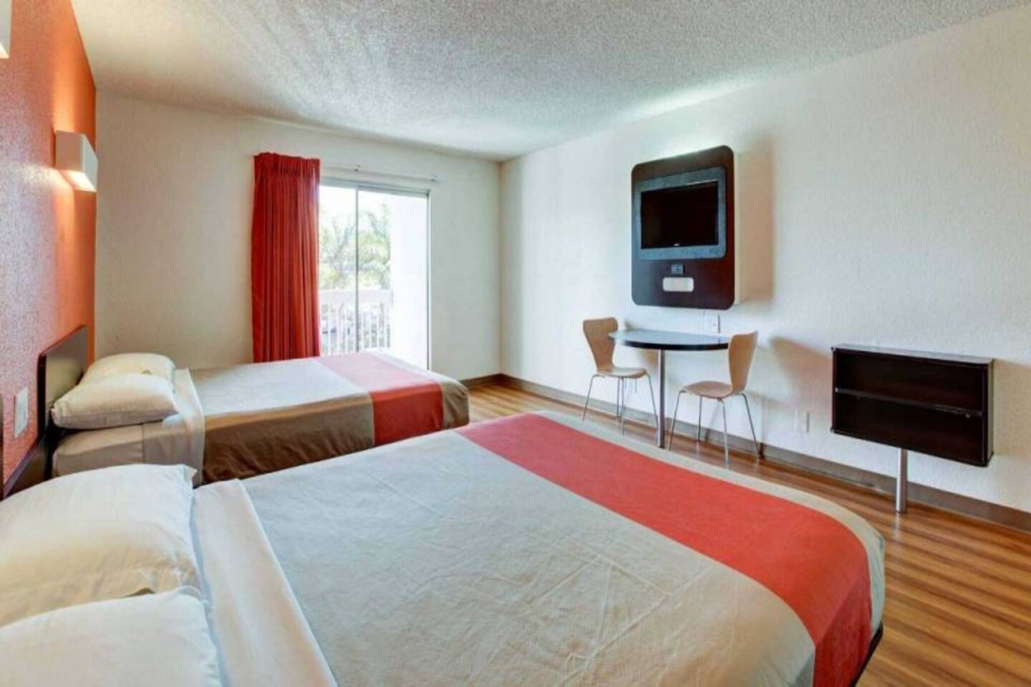A hotel room with two beds, a TV, a desk with chairs, and a window with curtains.