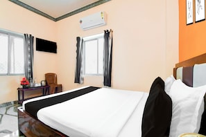 Ein Hotelzimmer mit Bett, Wandfernseher, Fenster mit Vorhängen, einem kleinen Tisch mit Stuhl und einem dekorativen Gegenstand auf dem Tisch.