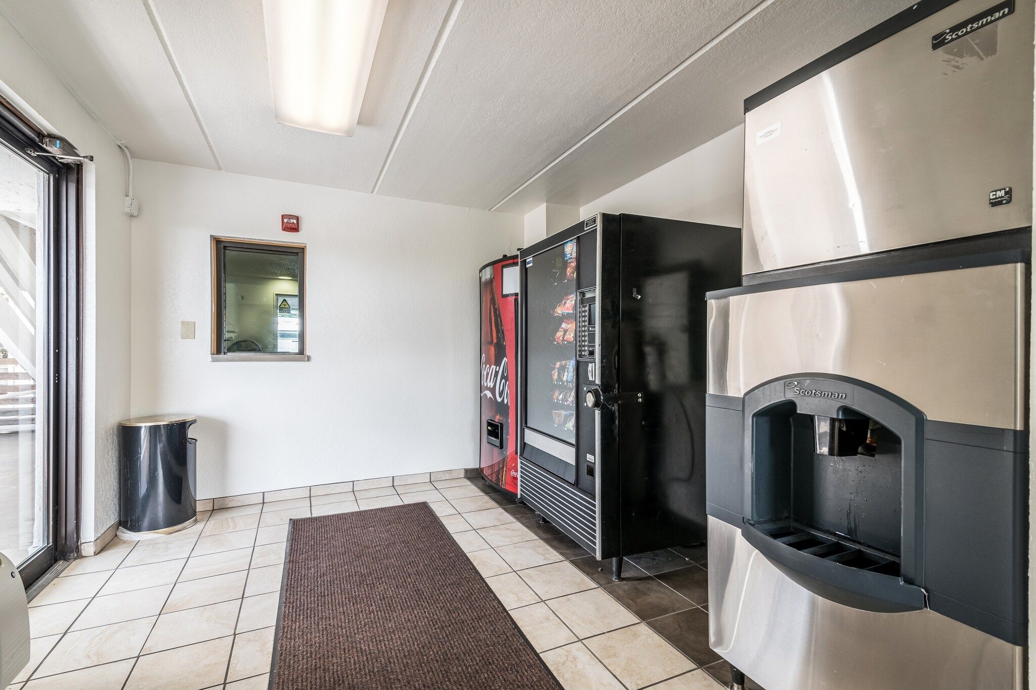A hallway with a vending machine, a trash bin, a fire alarm, and a Scotsman refrigerator.