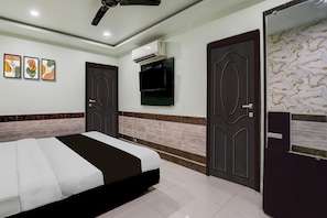 Habitación de hotel con una cama, ventilador de techo, equipo de aire acondicionado, televisor de pantalla plana y una obra de arte en la pared.