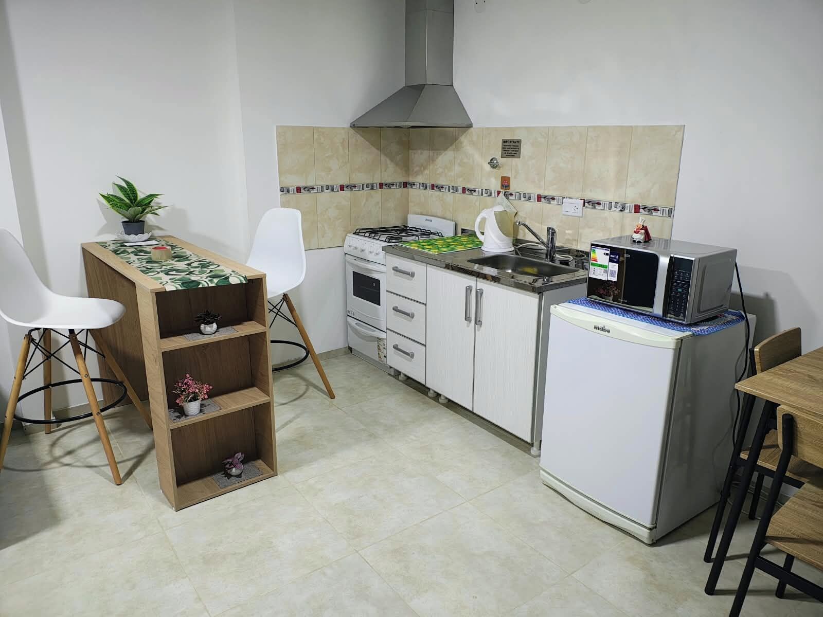 Cozinha compacta com fogão a gás, micro-ondas e pequena geladeira. Há uma cadeira branca e uma mesa de madeira com uma planta em cima.