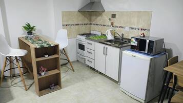 Cozinha compacta com fogão a gás, micro-ondas e pequena geladeira. Há uma cadeira branca e uma mesa de madeira com uma planta em cima.