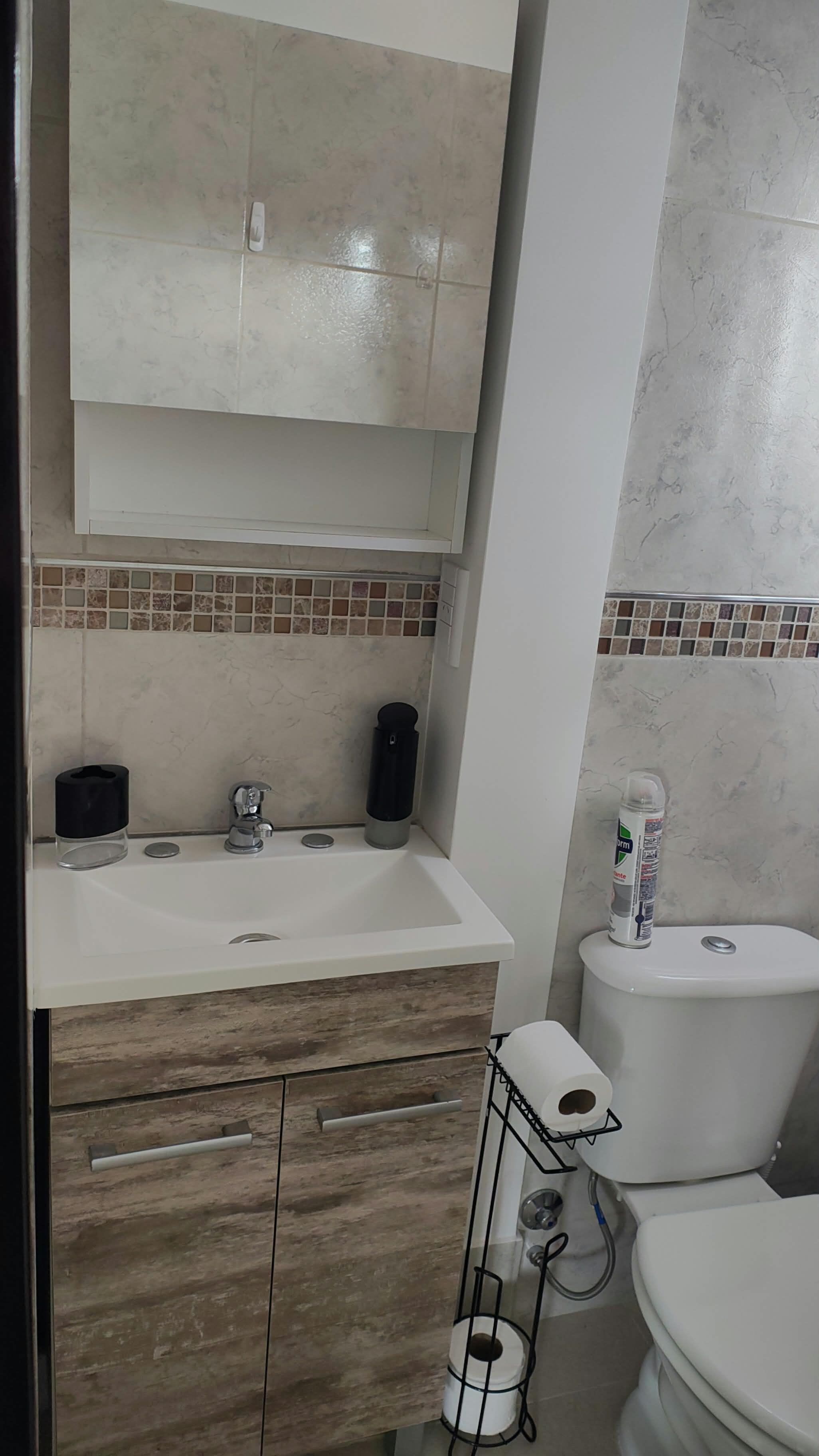 Casa de banho com lavatório branco, toucador em madeira, sanita e painel de azulejos.