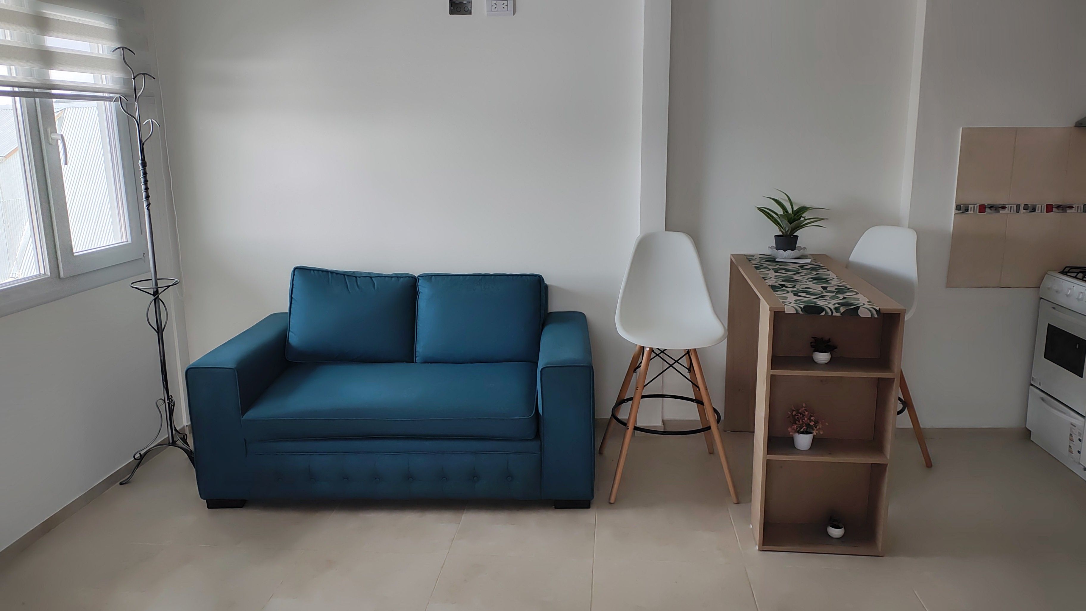 A room with a blue sofa, a white chair, a wooden side table with a potted plant, and a kitchenette area.